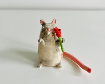 May include: A ceramic figurine of a gray rat holding a red flower with a green stem. The rat has a pink tail and ears. The figurine is on a white surface.