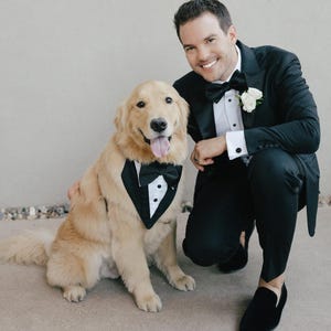 以下が含まれることがあります： 黒いタキシードのバンダナと蝶ネクタイを着けたゴールデンレトリバー犬が、白い花のブートニアが付いた黒いタキシードを着た人の隣に座っています。犬と人は微笑んでいます。