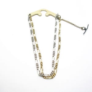 May include: A gold-toned tie clip with two parallel chains, one gold and one silver, connected to a T-bar. The clip has a decorative, curved design. The chains are made of a repeating link pattern.