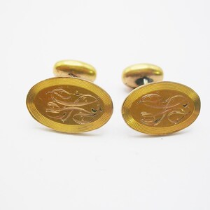 Puede incluir: Un par de gemelos dorados. Cada gemelo presenta una cara ovalada con un diseño grabado y una parte trasera redondeada. El diseño grabado parece ser un patrón floral o de desplazamiento abstracto. Los gemelos están sobre un fondo blanco.