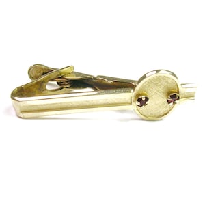 May include: A gold-toned tie clip with a rectangular base and a circular accent. The circular accent features two small, red gemstones. The clip is designed to secure a tie and add a touch of elegance.