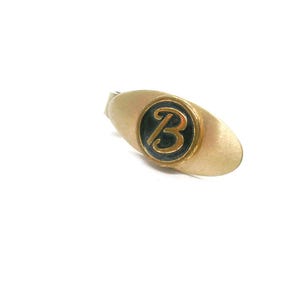 May include: A gold-toned tie clip with a black and gold circular emblem featuring the letter "B". The clip has a brushed metal finish and an oval shape. The design is simple and elegant.