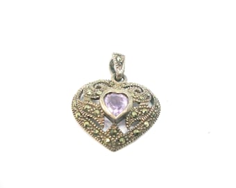 Può includere: Un ciondolo a forma di cuore in argento con una pietra preziosa viola al centro. Il ciondolo è decorato con un design a filigrana e piccole pietre grigio scuro.