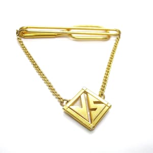 May include: A gold-coloured tie clip with a chain and a diamond-shaped pendant. The pendant features a geometric design with the letters "JS" in a stylized font. The tie clip is on a white background.