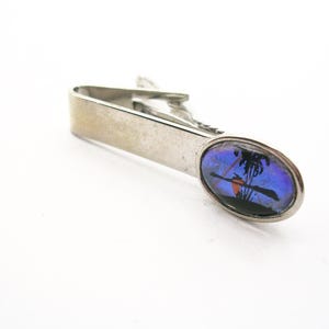May include: Silver tie clip with an oval inset depicting a tropical scene with a palm tree, boat, and sunset. The inset has a blue and purple background. The clip is made of metal.