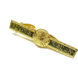 Puede incluir: Un pasador de corbata dorado con las palabras "LYONS FALLS" grabadas. El pasador presenta un emblema circular en el centro. El diseño es vintage y probablemente de metal.