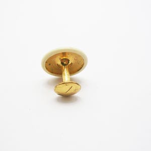 Pode incluir: Um único botão de punho dourado com uma face circular creme. O botão de punho tem um pino e uma pequena parte traseira redonda. O design é simples e elegante, adequado para roupas formais.