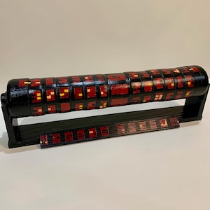 May include: A black wooden cylinder with a red and yellow geometric pattern is displayed on a black wooden stand. The cylinder is made of multiple rings that can be rotated.