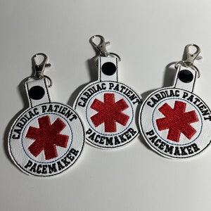 May include: Three white keychains with the text "CARDIAC PATIENT PACEMAKER" embroidered in black. Each keychain features a red star of life symbol. The keychains have silver-colored clasps.