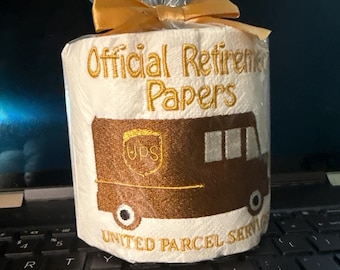 Puede incluir: Un rollo de papel higiénico con las palabras "Official Retirement Papers" bordadas en oro. Un camión UPS marrón está bordado debajo del texto. El rollo está envuelto en celofán y atado con un lazo dorado.