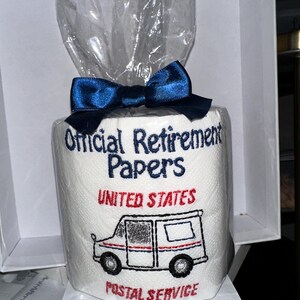 Pu&ograve; includere: Un rotolo di carta igienica bianca con le parole "Official Retirement Papers" ricamate in blu. Le parole "United States Postal Service" e un camion postale sono ricamate in rosso e nero. Il rotolo &egrave; avvolto in cellophane e legato con un fiocco blu.