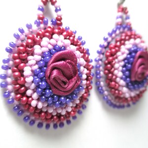 May include: Handmade beaded earrings in shades of purple, pink, and red. Each earring features a central fabric rose in a deep burgundy color. The earrings are round and teardrop-shaped, showcasing intricate beadwork.