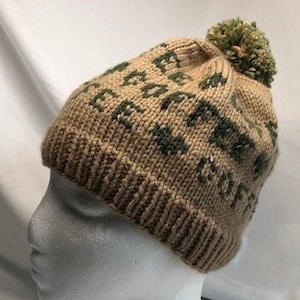 May include: A knitted beanie hat in tan with a green geometric pattern and a green pom-pom on top. The hat is displayed on a white mannequin head, showcasing its design and texture. The hat is a winter accessory.