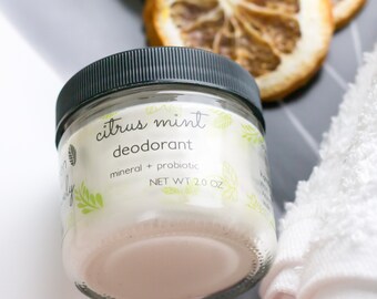 May include: A glass jar of "Citrus Mint Deodorant" with a black lid, next to a white towel and dried citrus slices. The label reads "mineral + probiotic" and "NET WT 2.0 OZ".