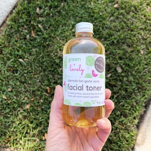 May include: A clear glass bottle of facial toner with a silver cap, held against a backdrop of green grass. The label reads "green + lovely" and "blemish-be-gone acne facial toner." The bottle contains 9 fl oz of product.