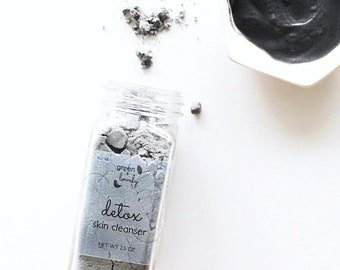May include: A glass jar containing grey powder, labelled "detox skin cleanser", sits beside a small white bowl filled with a dark grey paste. The jar's label reads "green + lovely" and "NET WT 2.5 OZ". Grey powder is scattered on the white surface.