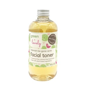 May include: A clear glass bottle of "green + lovely" facial toner. The label features green floral designs, a pink bird, and the text "blemish-be-gone acne facial toner." The bottle contains a yellow liquid and has a silver cap. It is 9 fl oz.