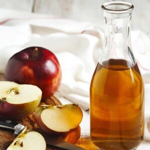 May include: A clear glass bottle filled with amber liquid, next to a whole red apple and apple slices on a wooden cutting board. A black knife rests on the board. The background includes a white cloth.