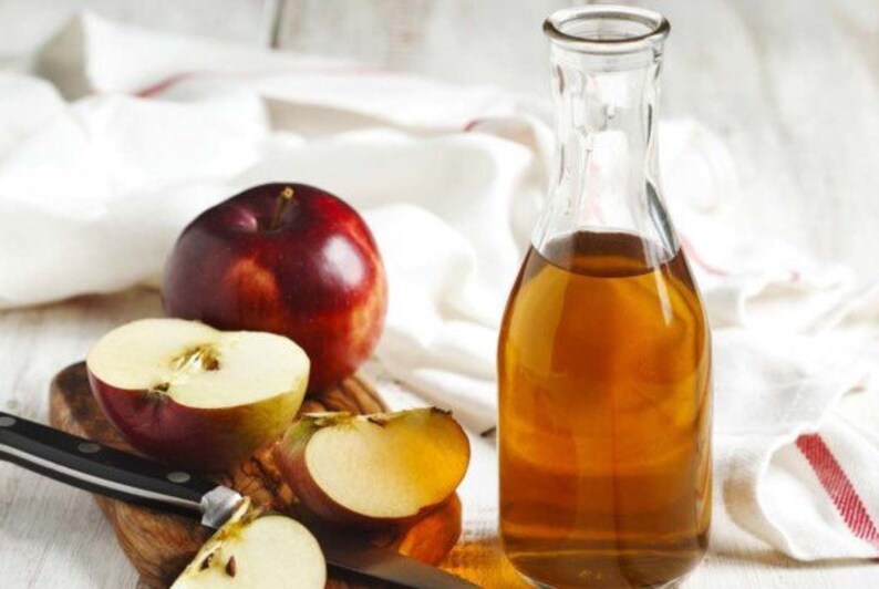 May include: A clear glass bottle filled with amber liquid, next to a whole red apple and apple slices on a wooden cutting board. A black knife rests on the board. The background includes a white cloth.