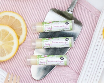 May include: Three lip balm tubes with the text "green lovely lemon meringue" are arranged on a silver server. The lip balms are on a pink cloth with lemon slices and a wooden fork and knife. The lip balm is labeled "nature's silk lip balm".