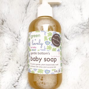 May include: A clear plastic bottle of baby soap with a white pump dispenser. The label reads "green + lovely for baby" and "gentle bottom's baby soap." The bottle contains a light brown liquid and is 266 ml.