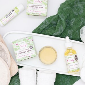 May include: An overhead shot of skincare products, including a lip balm, salve, face serum, and cotton pads. The products are labeled "calendula" and "Green Lovely." A green cloth and white tray are also visible.