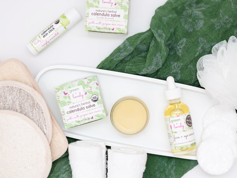 May include: An overhead shot of skincare products, including a lip balm, salve, face serum, and cotton pads. The products are labeled "calendula" and "Green Lovely." A green cloth and white tray are also visible.