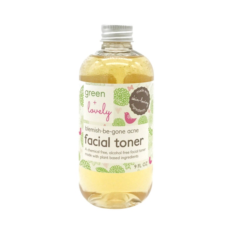 May include: A clear glass bottle of "green + lovely" facial toner. The label features green floral designs, a pink bird, and the text "blemish-be-gone acne facial toner." The bottle contains a yellow liquid and has a silver cap. It is 9 fl oz.