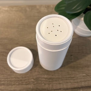 Peut inclure: Un récipient cylindrique blanc avec un couvercle amovible. Le dessus du récipient présente un motif perforé. Le couvercle est sur le côté. Le récipient est sur une surface en bois.
