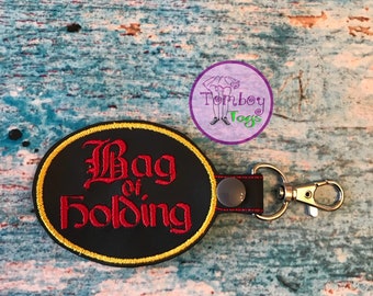 May include: Black oval keychain with yellow stitching and a red embroidered design that says "Bag of Holding".