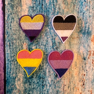 May include: Four heart-shaped patches with embroidered edges. Each patch features a different color scheme representing various LGBTQ+ identities. The patches are yellow, white, purple, black, pink, blue, and brown. The patches are on a blue and green textured surface.