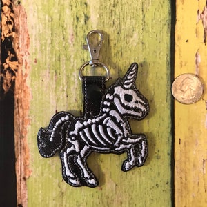 May include: A black and white embroidered skeleton unicorn keychain with a silver clip. The keychain is about the size of a quarter.