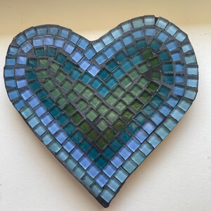May include: A heart-shaped mosaic wall decoration constructed from small, square glass tiles in various shades of blue, teal, and green. The tiles are arranged in concentric layers, transitioning from light blue on the outer edge to deep green.