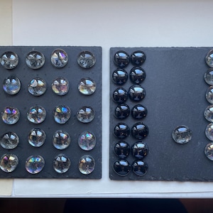 May include: Two rectangular black slate boards arranged side by side. The left board features twenty-five clear, iridescent glass pebbles in a five-by-five grid. The right board displays sixteen solid black glass pebbles and nine clear glass pebbles.