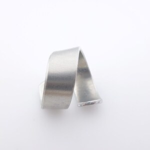 K&ouml;nnte beinhalten: Ein Ring aus geb&uuml;rstetem Silbermetall mit einem geschwungenen, bandartigen Design. Der Ring hat eine glatte, matte Oberfl&auml;che und eine schlichte, moderne &Auml;sthetik. Der Ring wird vor einem wei&szlig;en Hintergrund gezeigt.