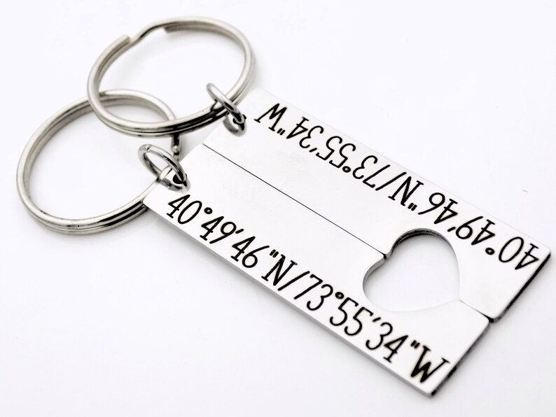 May include: A silver keychain with two key rings and a rectangular tag. The tag is split into two sections, each with a set of coordinates engraved on it. The coordinates are 40&deg;49&prime;46&Prime;N/73&deg;55&prime;34&Prime;W. The tag has a heart-shaped cutout in the middle.