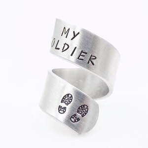 K&ouml;nnte beinhalten: Ein silberfarbener Wickelring mit den Worten "MY SOLDIER" auf dem oberen Band. Das untere Band zeigt zwei kleine Fu&szlig;abdruck-Designs. Der Ring besteht aus einem gl&auml;nzenden, metallischen Material.