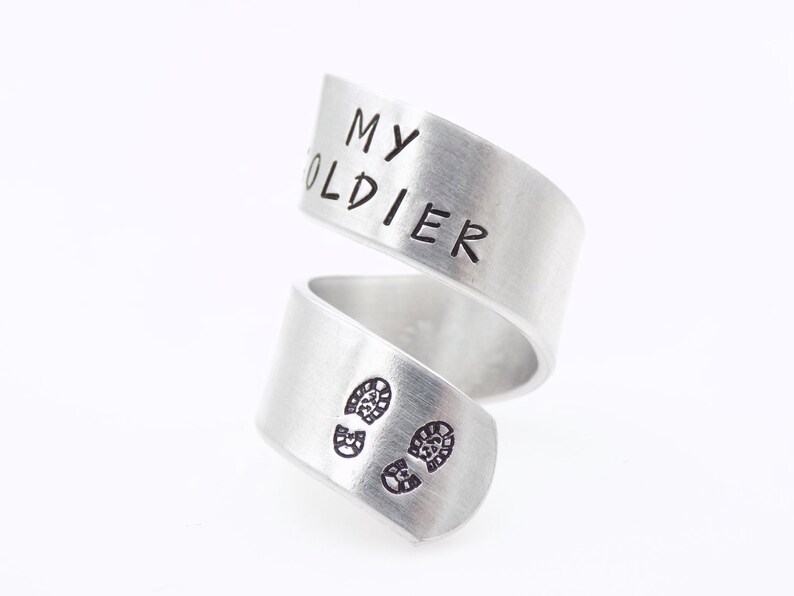 K&ouml;nnte beinhalten: Ein silberfarbener Wickelring mit den Worten "MY SOLDIER" auf dem oberen Band. Das untere Band zeigt zwei kleine Fu&szlig;abdruck-Designs. Der Ring besteht aus einem gl&auml;nzenden, metallischen Material.
