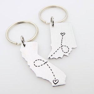 May include: Two silver keychains featuring the outlines of Indiana and California. A dotted line with hearts connects the two states.