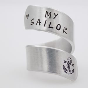 K&ouml;nnte beinhalten: Ein silberfarbener Wickelring mit den Worten "MY SAILOR" und einem Herzsymbol, das auf das obere Band graviert ist. Das untere Band zeigt ein Ankersymbol. Der Ring hat ein geb&uuml;rstetes Metall-Finish.