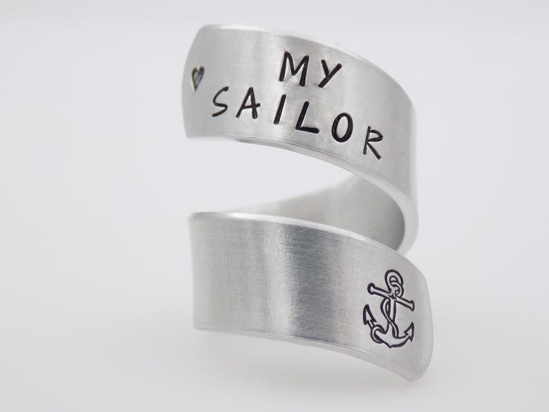 K&ouml;nnte beinhalten: Ein silberfarbener Wickelring mit den Worten "MY SAILOR" und einem Herzsymbol, das auf das obere Band graviert ist. Das untere Band zeigt ein Ankersymbol. Der Ring hat ein geb&uuml;rstetes Metall-Finish.