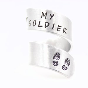 K&ouml;nnte beinhalten: Ein silberfarbener Wickelring mit den Worten "MY SOLDIER" auf dem oberen Band eingraviert. Das untere Band zeigt zwei kleine, gestempelte Fu&szlig;abdr&uuml;cke. Der Ring ist vor wei&szlig;em Hintergrund.