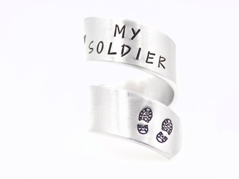 K&ouml;nnte beinhalten: Ein silberfarbener Wickelring mit den Worten "MY SOLDIER" auf dem oberen Band eingraviert. Das untere Band zeigt zwei kleine, gestempelte Fu&szlig;abdr&uuml;cke. Der Ring ist vor wei&szlig;em Hintergrund.