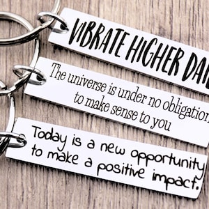 May include: Three silver keychains with inspirational quotes. The top keychain says "Vibrate Higher Daily." The middle keychain says "The universe is under no obligation to make sense to you." The bottom keychain says "Today is a new opportunity to make a positive impact."