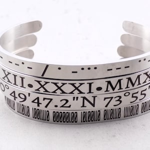 May include: A silver metal bracelet with four bands. The bands are engraved with Roman numerals, a date, and coordinates. The coordinates are 49' 47.2"N 73° 55' 35"