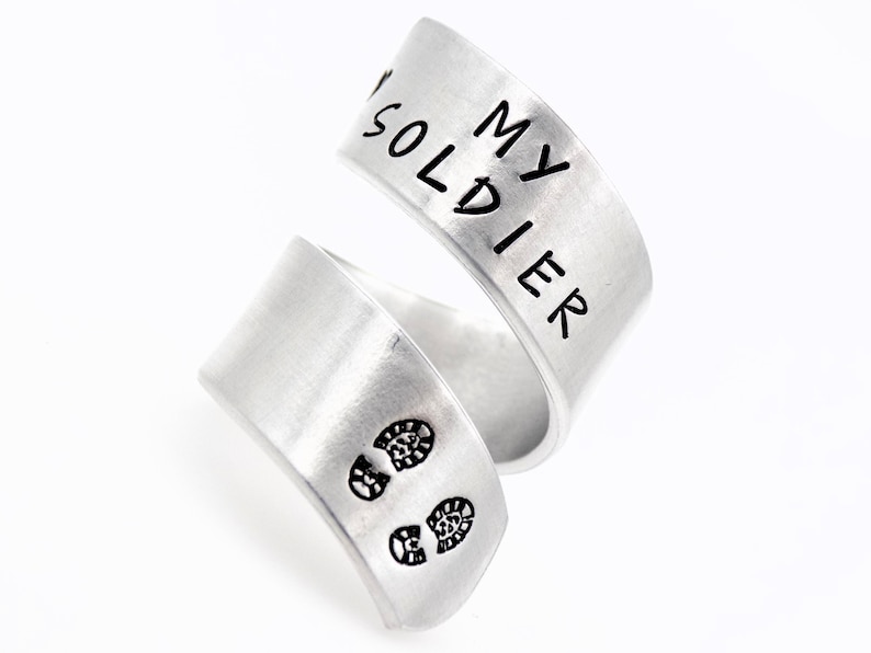 K&ouml;nnte beinhalten: Ein silberfarbener Wickelring mit der Aufschrift "MY SOLDIER" auf dem oberen Band. Das untere Band zeigt zwei kleine schwarze Fu&szlig;abdr&uuml;cke. Der Ring ist in einem einzigartigen, &uuml;berlappenden Stil gestaltet.