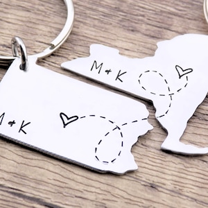 May include: Two silver keychains shaped like states with a dotted line connecting them. The keychains are engraved with the initials "M+K" and a heart.