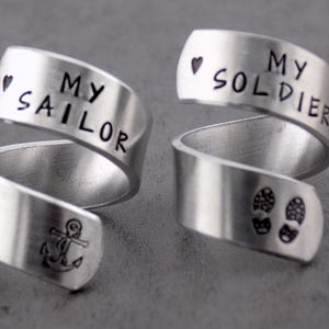K&ouml;nnte beinhalten: Zwei silberfarbene Wickelringe. Ein Ring hat die Worte "MY SAILOR" mit einem Ankersymbol und einem Herz. Der andere Ring hat die Worte "MY SOLDIER" mit einem Herz und Fu&szlig;abdr&uuml;cken. Schmuck.