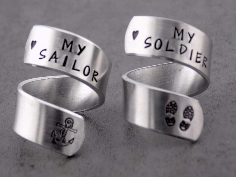 K&ouml;nnte beinhalten: Zwei silberfarbene Wickelringe. Ein Ring hat die Worte "MY SAILOR" mit einem Ankersymbol und einem Herz. Der andere Ring hat die Worte "MY SOLDIER" mit einem Herz und Fu&szlig;abdr&uuml;cken. Schmuck.