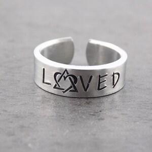 May include: A silver ring with the word "LOVED" engraved on it. The "O" is replaced with a symbol of two intertwined hearts.
